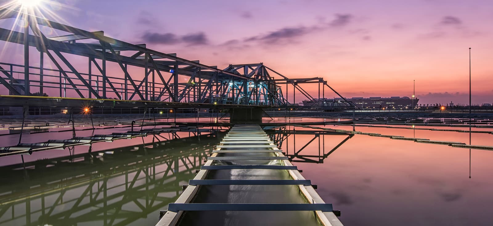 A wastewater clarifier bridge at dusk, its walkway reflected in still water